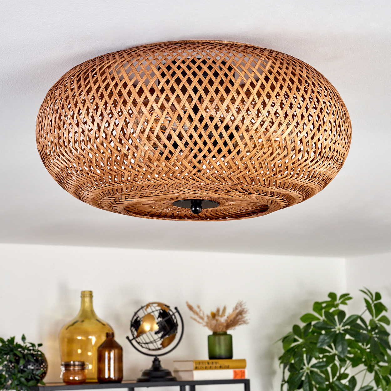 Eine gewebte Deckenlampe aus Rattan hängt über einem Regal mit Dekoration, darunter ein Globus, Vasen, Pflanzen und Bücher. Die Decke ist weiß.