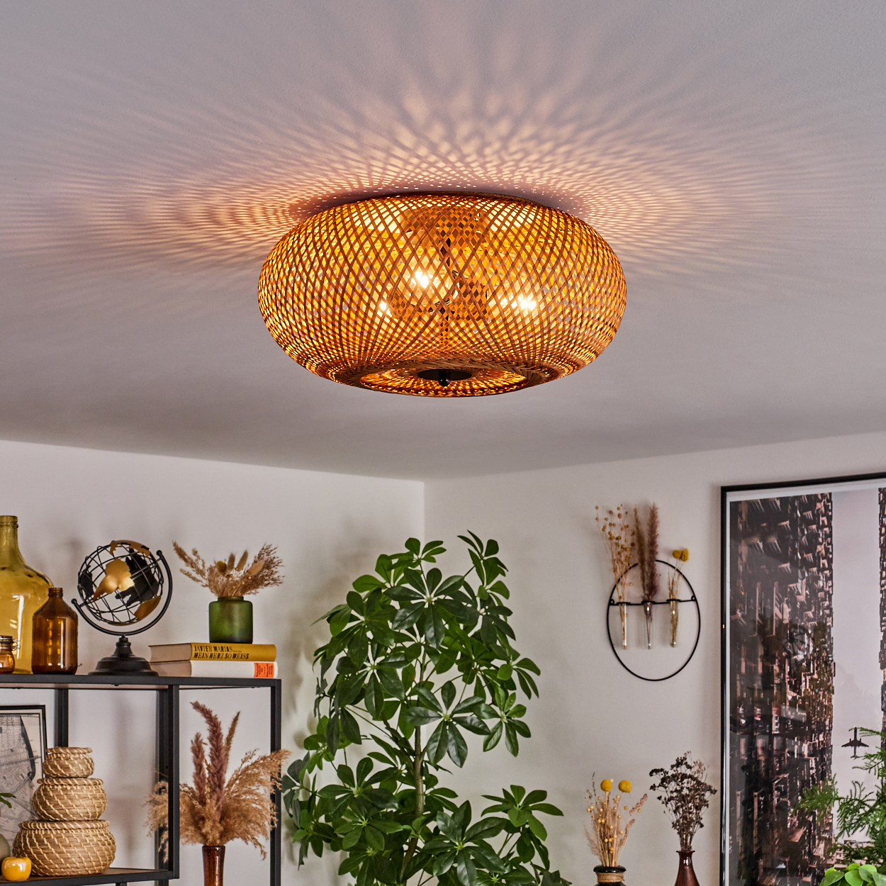 Eine Deckenlampe aus geflochtenem Rattan ist beleuchtet. Darunter ein Regal mit Deko und eine Blühpflanze. Im Hintergrund hängt ein Stadtfoto.