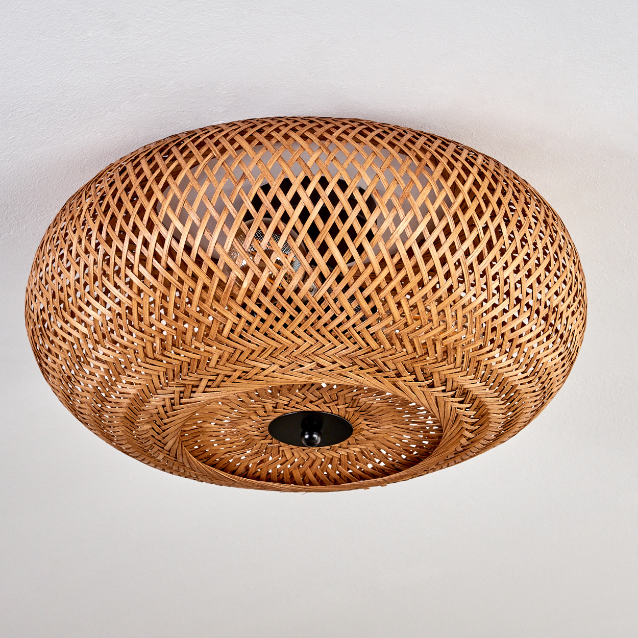 Eine gewebte Deckenlampe aus Rattan. Sie ist rund, braun und hat ein schwarzes Zentralelement. Der Hintergrund ist eine weiße Decke.