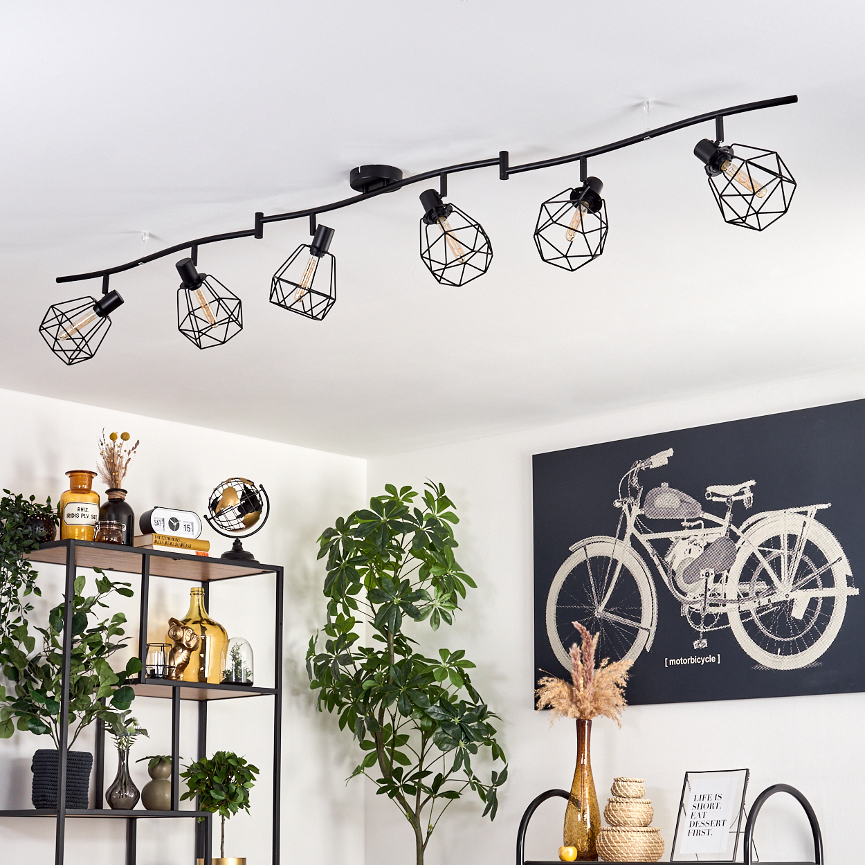 Ein modernes Wohnzimmer mit schwarzer Deckenleuchte. Ein schwarzes Regal mit Dekoration und ein Fahrradabdruck an der Wand.
