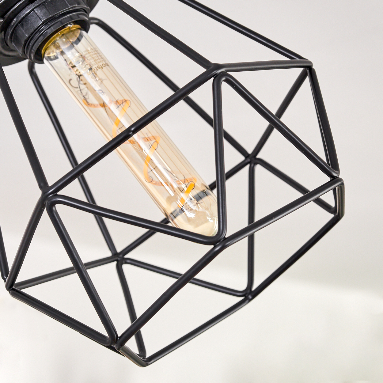 Nahaufnahme eines schwarzen, geometrischen Lampenschirms mit Edison-Glühbirne. Der Hintergrund ist eine weiße Oberfläche.