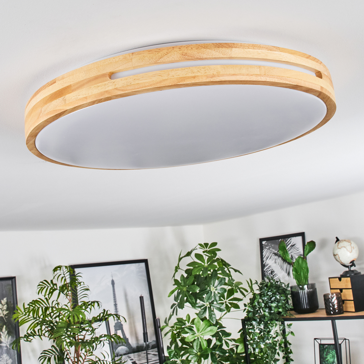 Eine Deckenlampe aus Holz und Weiß. Darunter sind Pflanzen, Bilder und ein Regal mit Dekoration zu sehen. Der Raum hat weiße Wände.