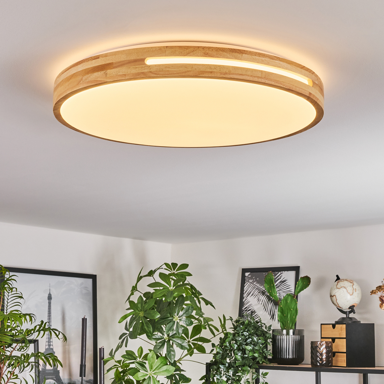 Eine kreisförmige Deckenlampe aus Holz beleuchtet einen Raum. Pflanzen und Bilder sind darunter sichtbar. Die Lampe ist rund, und das Licht ist warm.