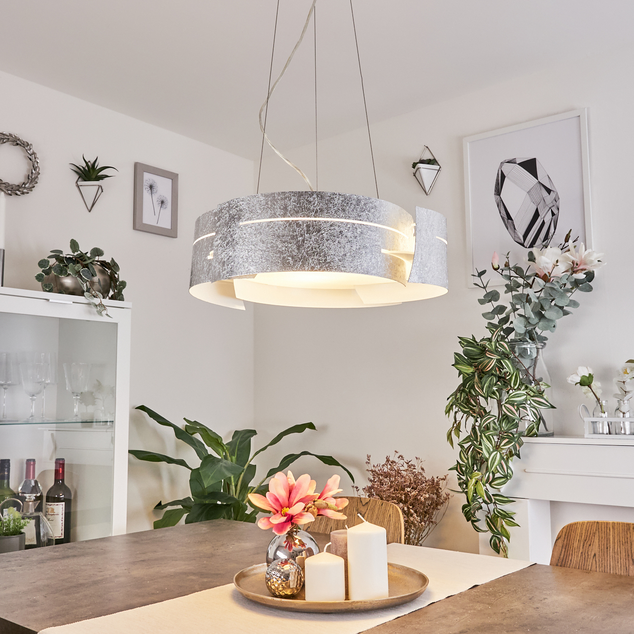 Ein Esszimmer mit einem großen grauen Tisch, einer dekorativen Lampe, Pflanzen und Kerzen auf dem Tisch. Es gibt einen weißen Schrank und Kunstwerke.