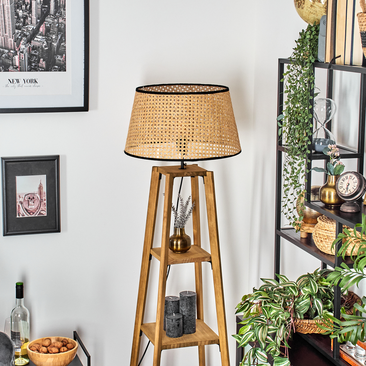 Eine hölzerne Stehlampe mit gewebtem Lampenschirm steht in einem Raum. Regale beherbergen Dekorationen, darunter Pflanzen und Kerzen. Bilder hängen an der Wand.