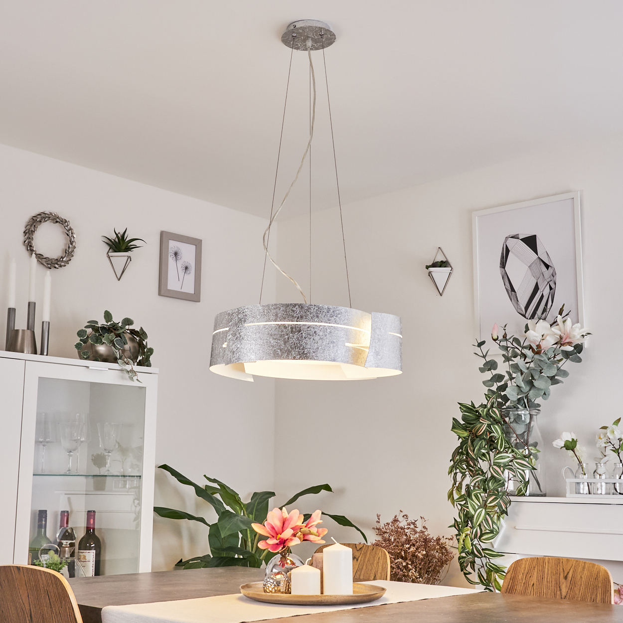 Ein modernes Esszimmer mit silberner Hängelampe, Tisch mit Kerzen und Pflanzen. Schrank und Kunstwerk im Hintergrund.