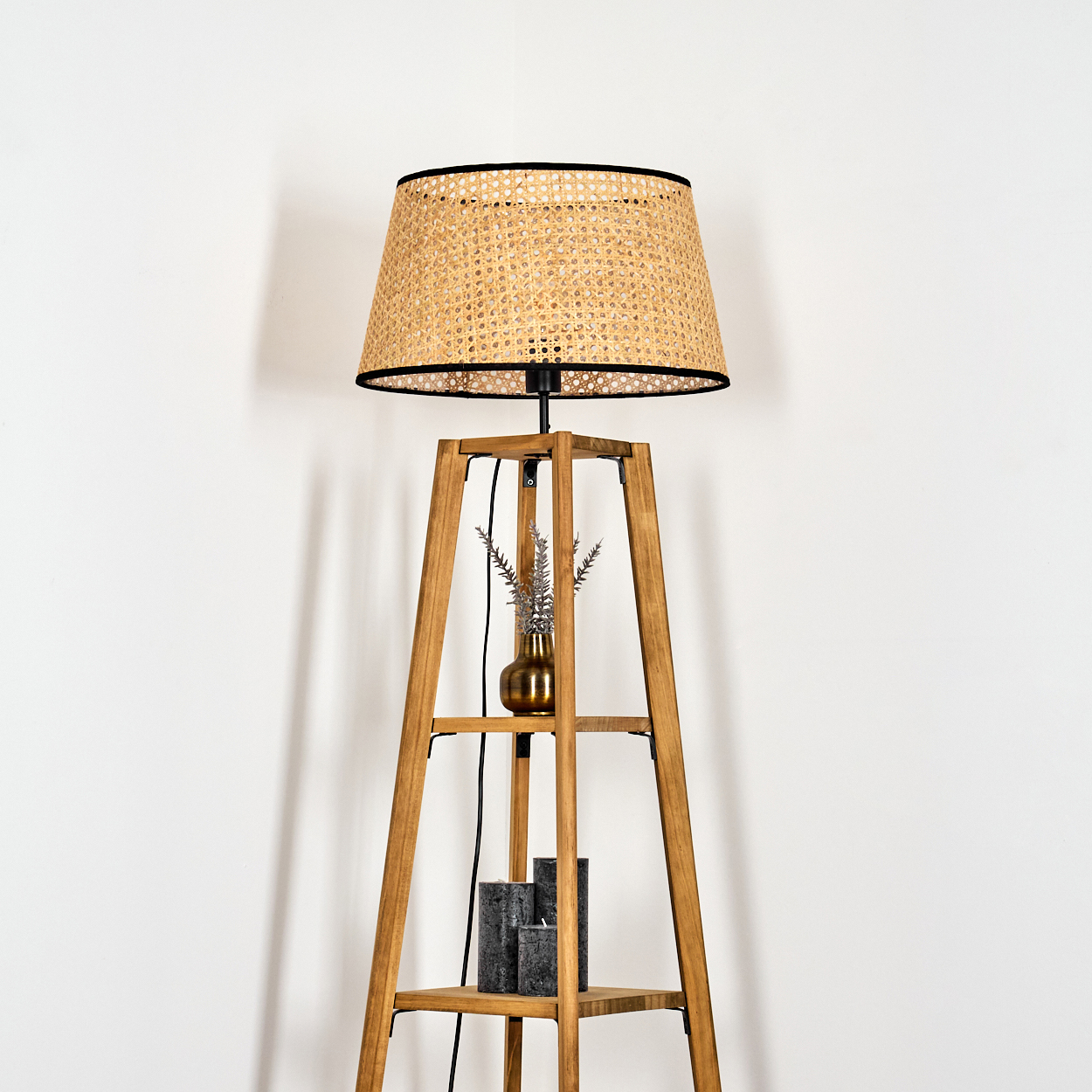 Eine hölzerne Stehlampe mit gewebtem Lampenschirm. Sie hat Regale mit einer Vase und Kerzen, vor einer weißen Wand.