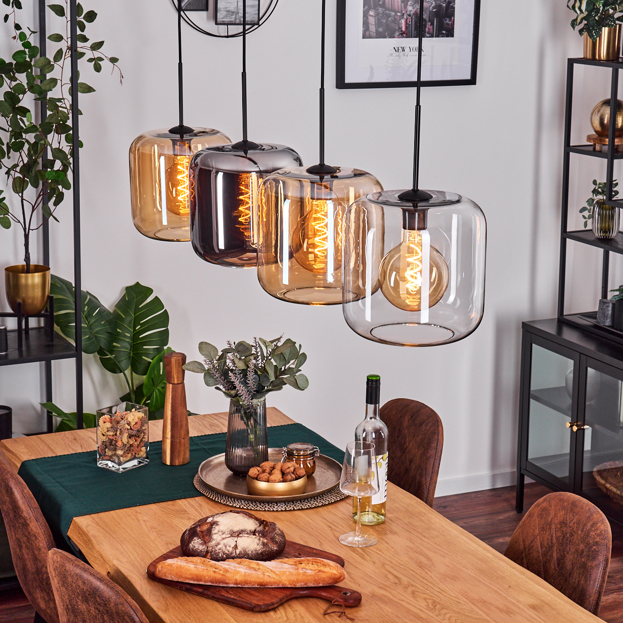 Szene im Esszimmer. Vier Pendelleuchten hängen über einem Holztisch mit Essen und Wein. Lederstühle umrunden den Tisch.