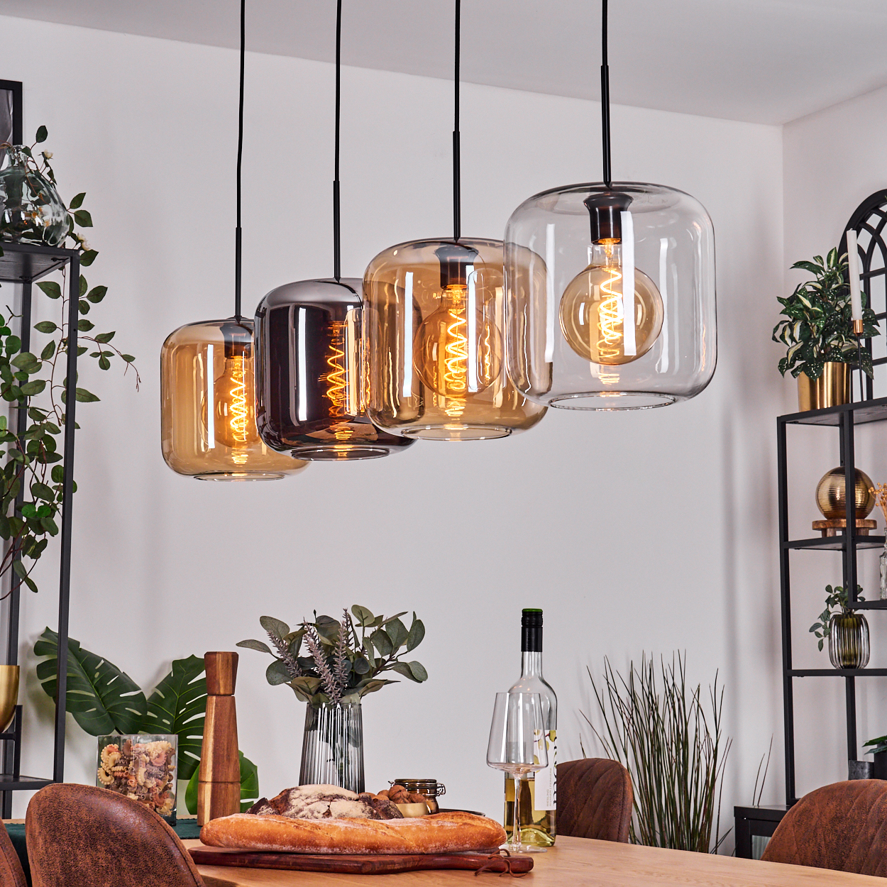 Esszimmer mit bernsteinfarbenen Hängelampen, Tisch mit Brot, Wein und Pflanzen. Schwarze Metallregale mit Dekoration.