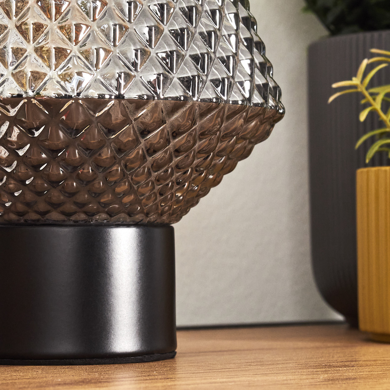 Nahaufnahme einer dekorativen Lampe mit schwarzem Sockel und strukturiertem, glasähnlichem Schirm. Pflanzen in Töpfen im Hintergrund.