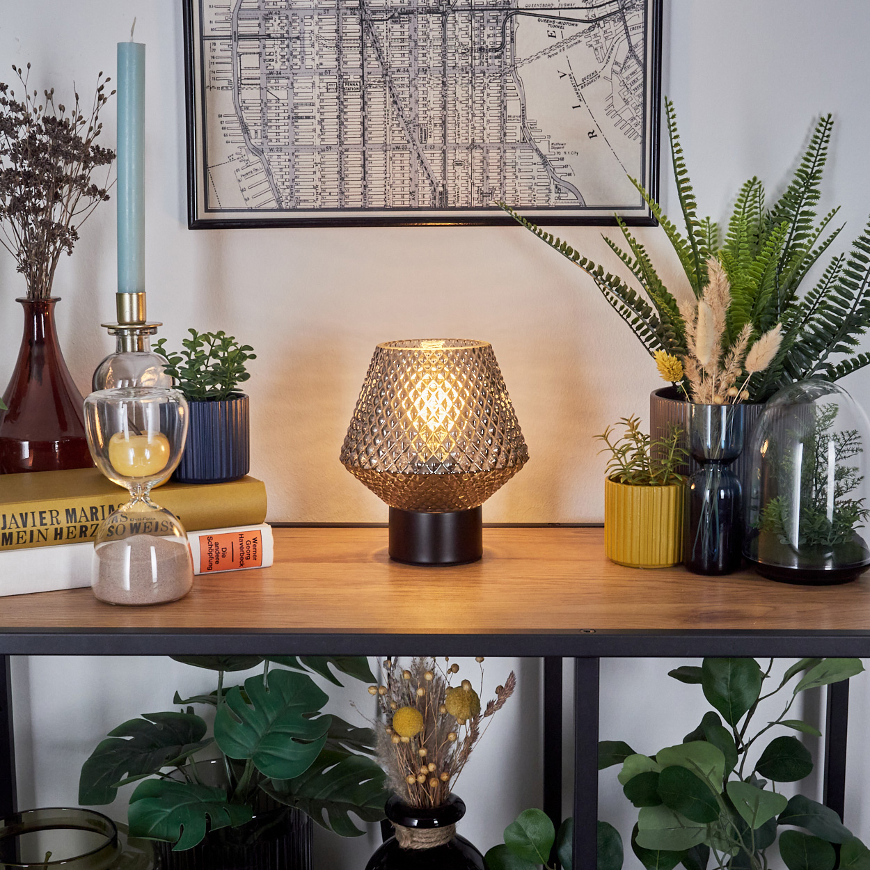Ein Tisch mit Lampe, Pflanzen, Büchern und einer Karte. Eine Kerze und eine Sanduhr sind ebenfalls vorhanden. Darunter ein Regal mit Pflanzen.