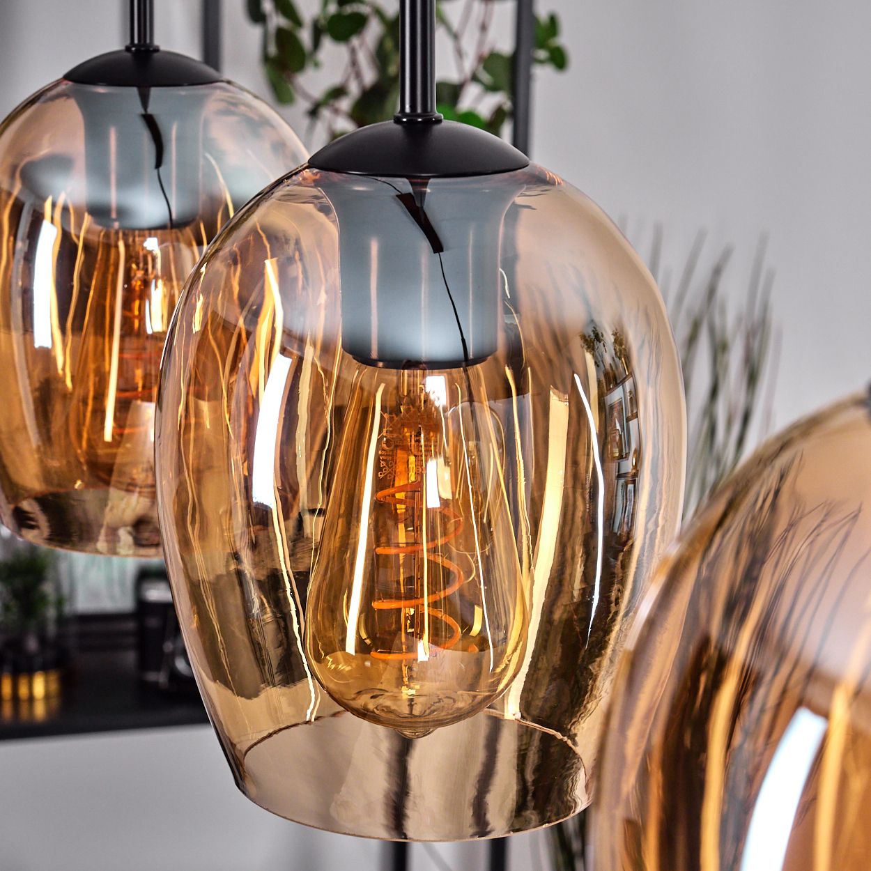 Nahaufnahme von drei bernsteinfarbenen Glas-Pendelleuchten mit freiliegenden Wendelglühbirnen. Schwarze Armaturen und Hintergrundlaub.