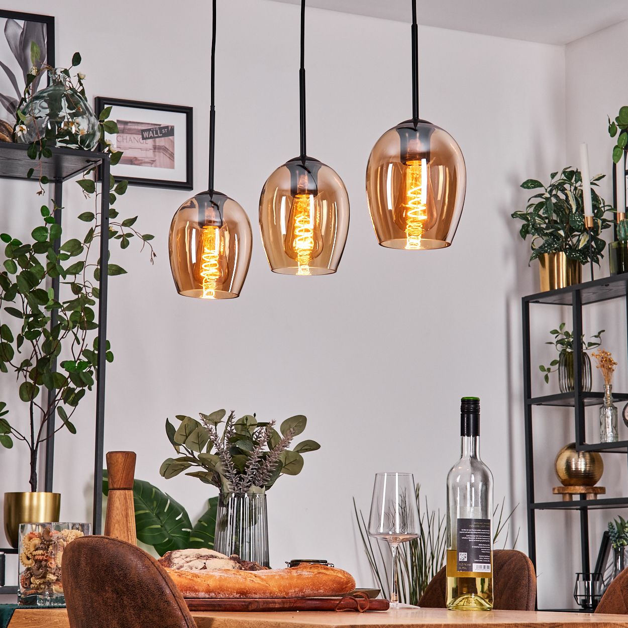 Esszimmer-Arrangement mit drei Pendelleuchten, Tisch mit Brot, Wein, Gläsern und dekorativen Pflanzen auf Regalen.