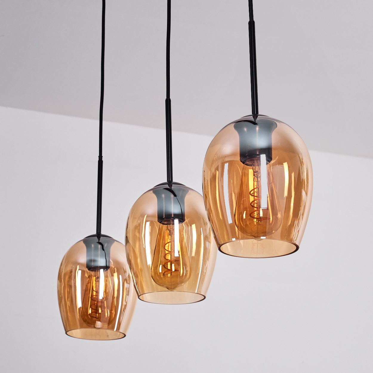 Drei moderne Pendelleuchten. Jede hat einen bernsteinfarbenen Glasschirm und eine sichtbare, glühende Glühbirne. Schwarze Kabel hängen von einer weißen Decke.