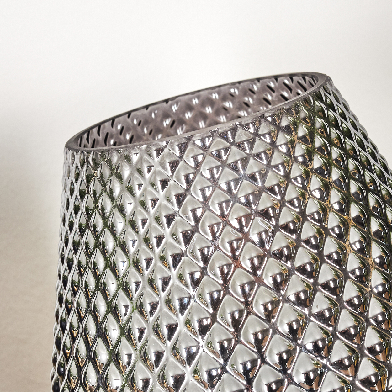 Eine geneigte, silberne Vase mit Rautenmuster. Der obere Teil hat Löcher. Der Hintergrund ist cremefarben.