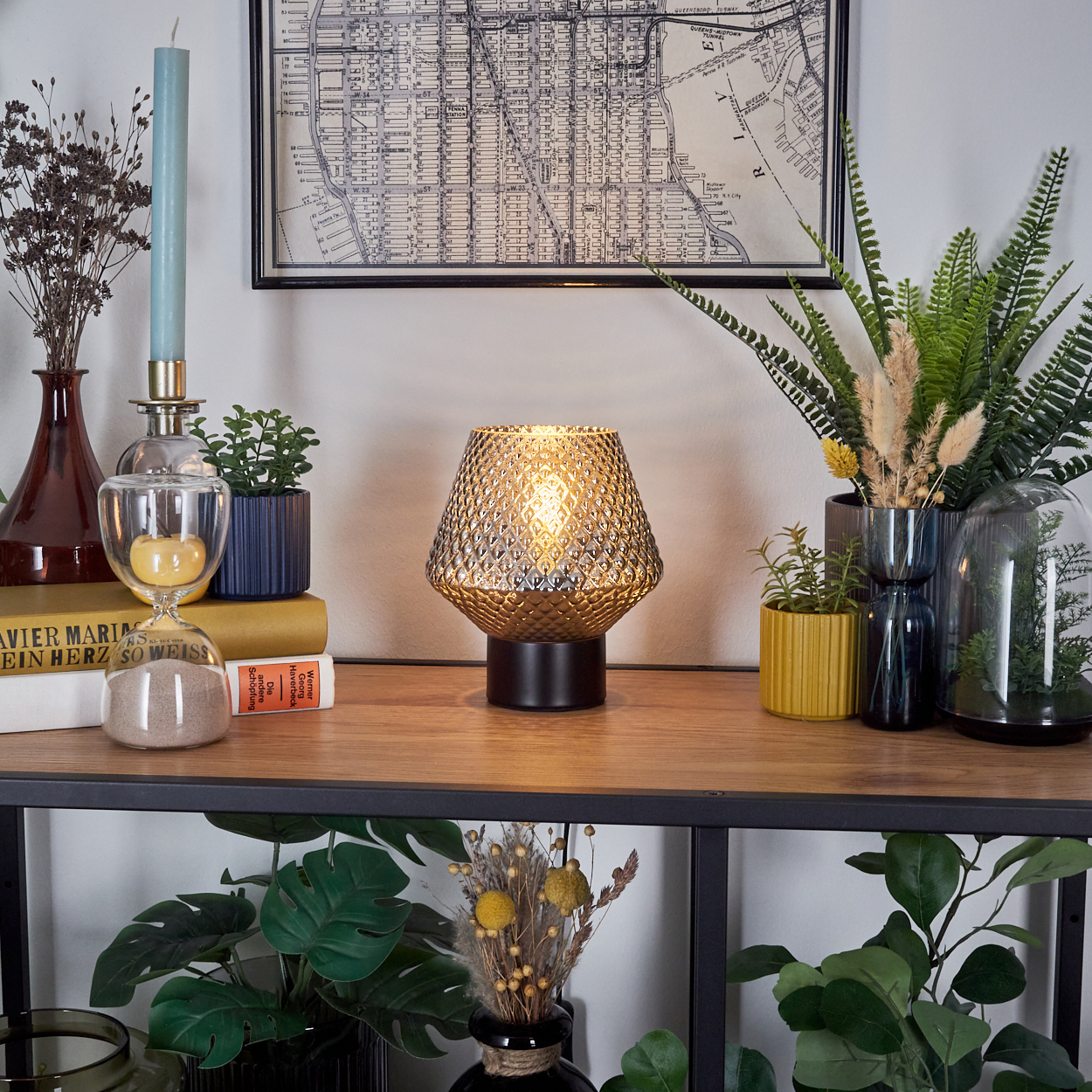 Ein dekoratives Regal mit Lampe, Pflanzen, Büchern und einer Karte. Die Lampe leuchtet und wirft einen warmen Schein.