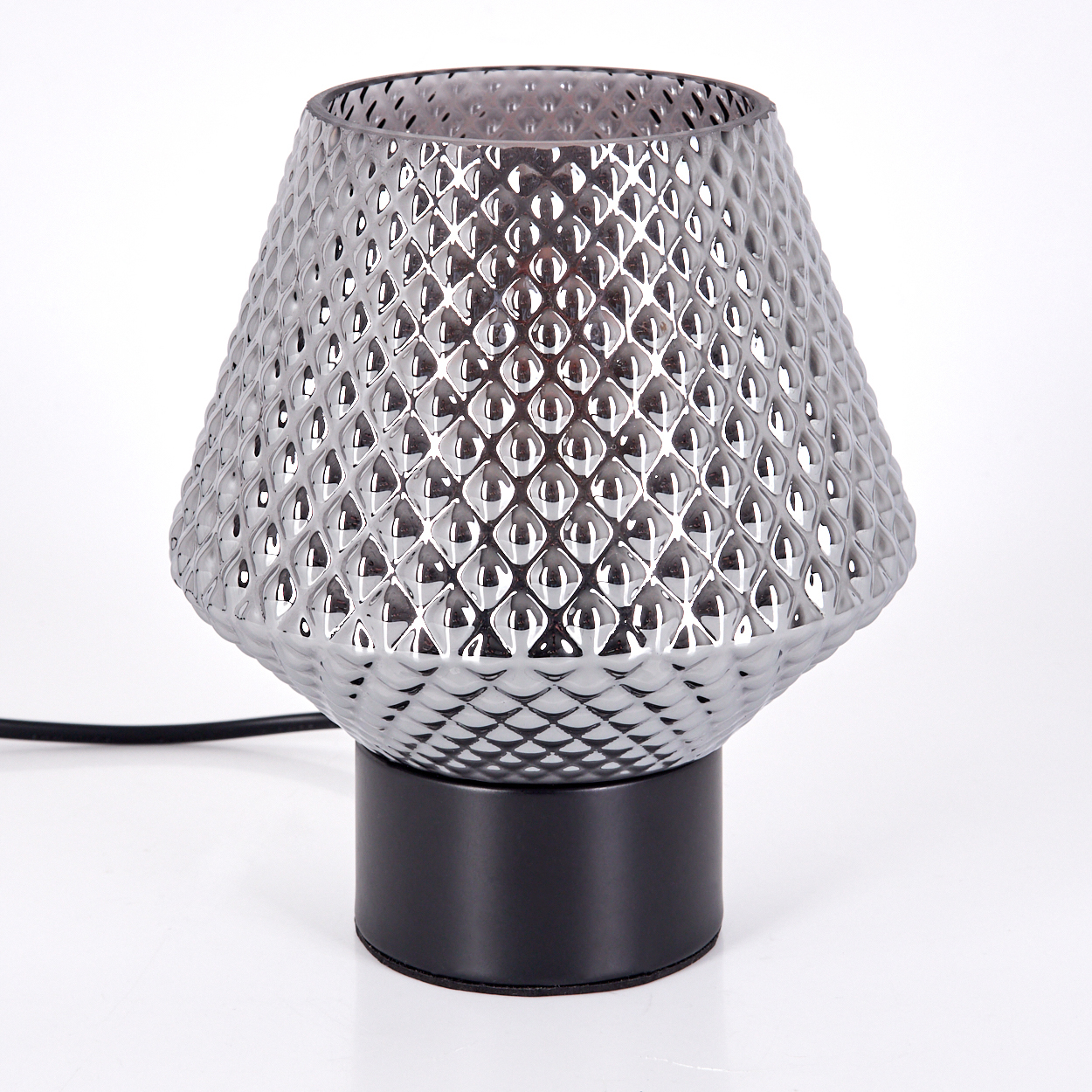 Eine silberne, strukturierte Lampe mit schwarzem Sockel steht auf einer weißen Oberfläche. Die Lampe hat einen gemusterten Schirm und einen zylindrischen Sockel.