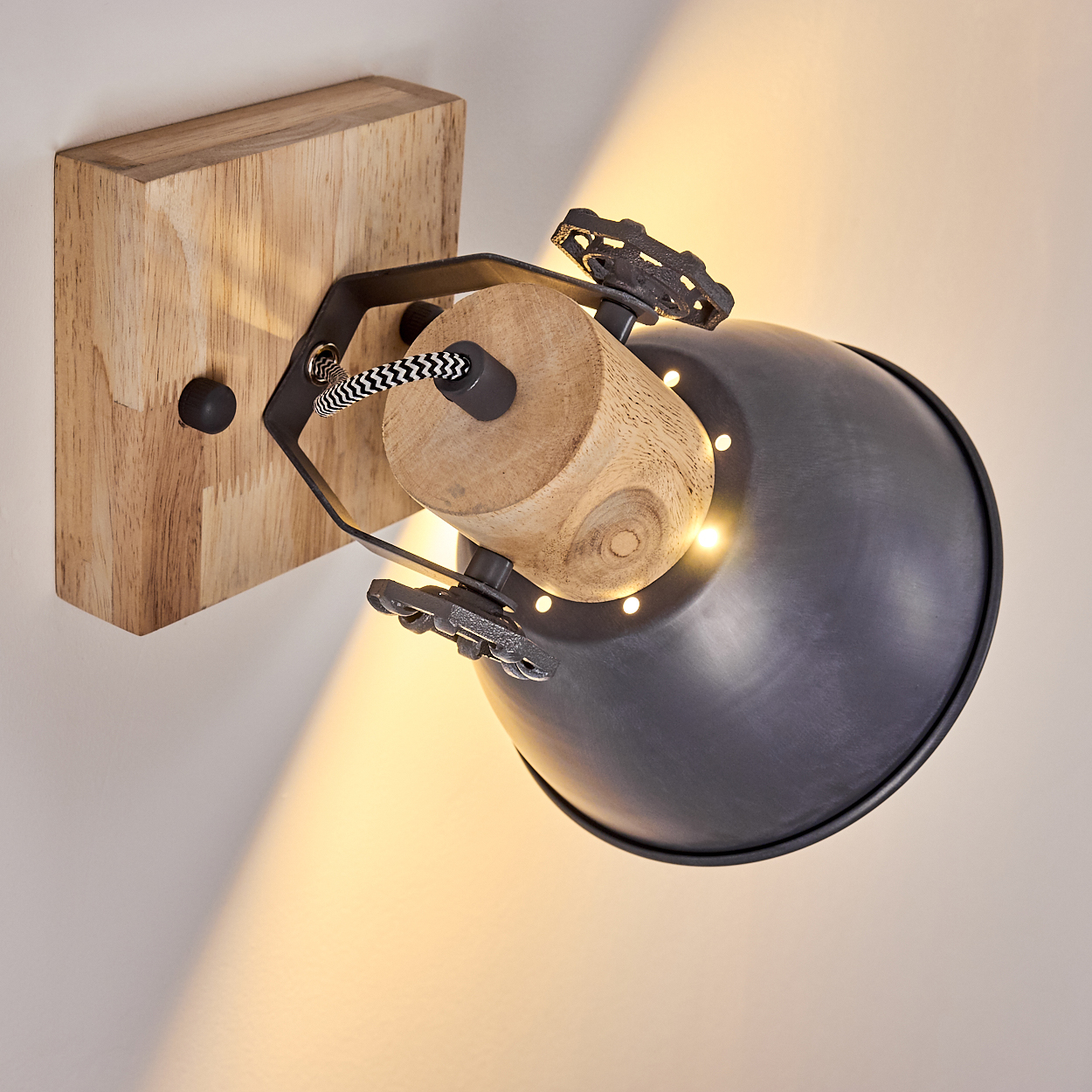 Eine Wandlampe mit Holzsockel, schwarzem Metallschirm und dekorativen Metalldetails. Das Licht scheint auf die Wand.