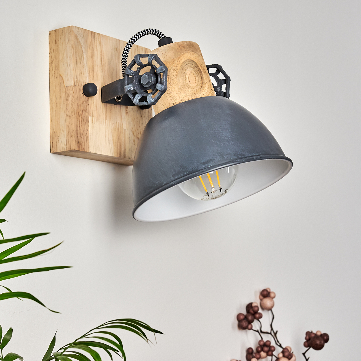 Eine Wandlampe mit Holzsockel, grauem Lampenschirm und sichtbarer Glühbirne. Dekorative Elemente sind eine Pflanze und ein Blumenarrangement.
