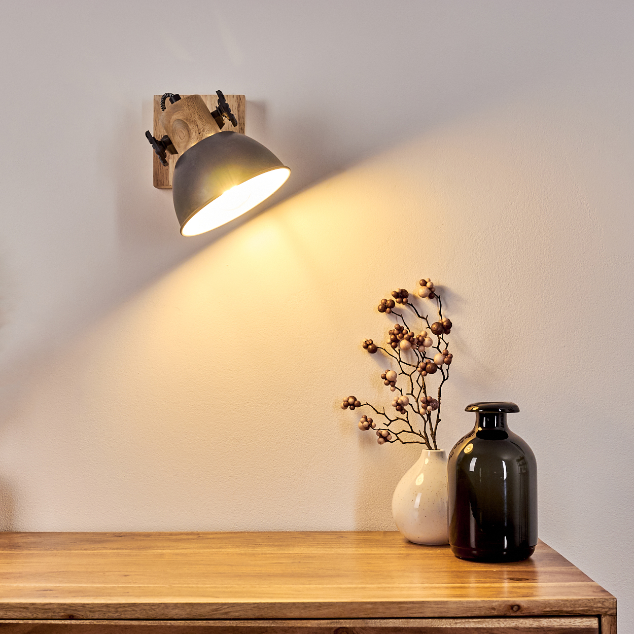 Eine Wandlampe scheint auf einen Holztisch mit Vasen und Blumen. Die Lampe ist auf einem Holzsockel montiert und beleuchtet die Wand.