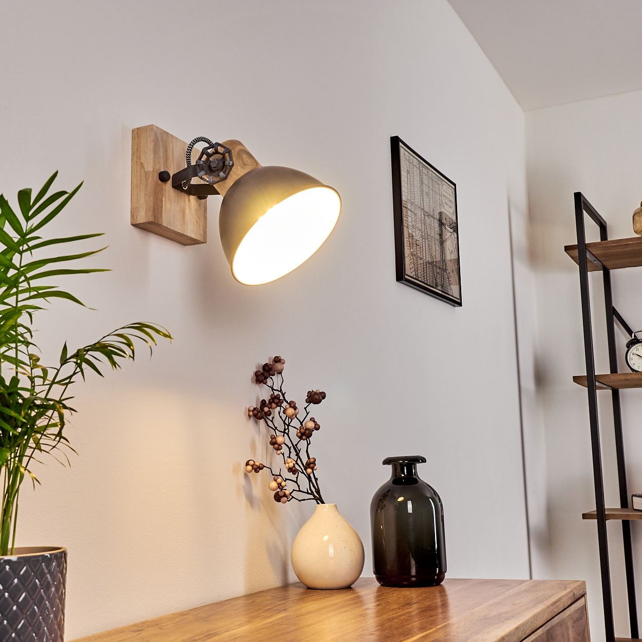 Eine Wandlampe mit Holzsockel leuchtet. Darunter stehen eine Vase mit Blumen und eine schwarze Vase auf einer Holzoberfläche.