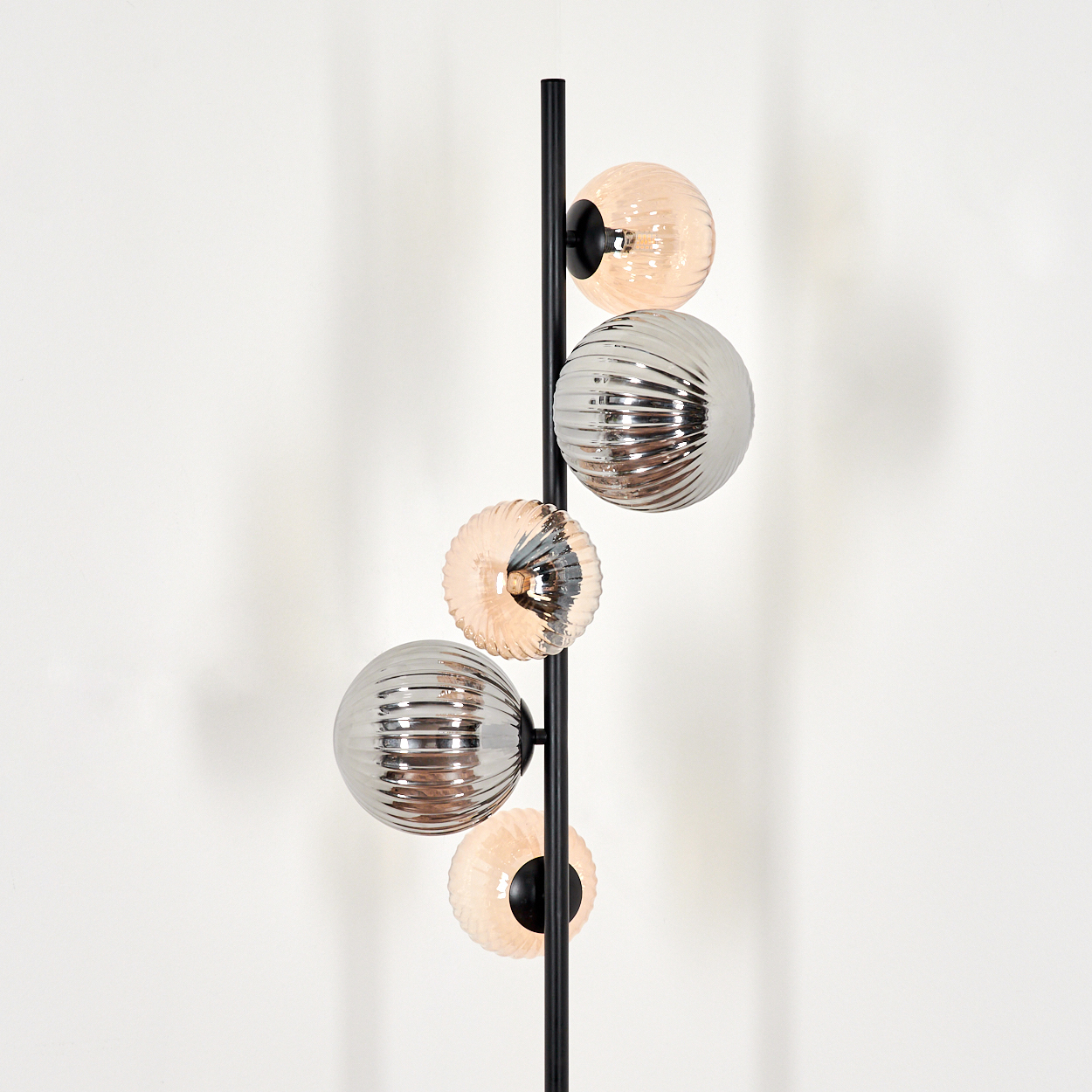 Eine schwarze Stehlampe mit fünf Glaskugeln. Die Kugeln sind geriffelt und wechseln sich zwischen Silber und Pfirsichfarben ab, vor einer weißen Wand.