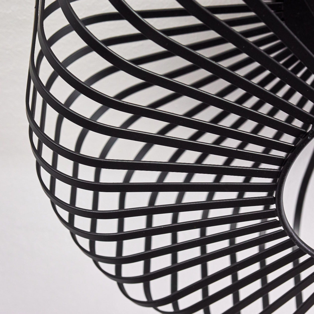 Nahaufnahme eines schwarzen Lampenschirms aus Drahtgeflecht vor weißem Hintergrund. Die Stäbe bilden ein spiralförmiges, käfigartiges Design.