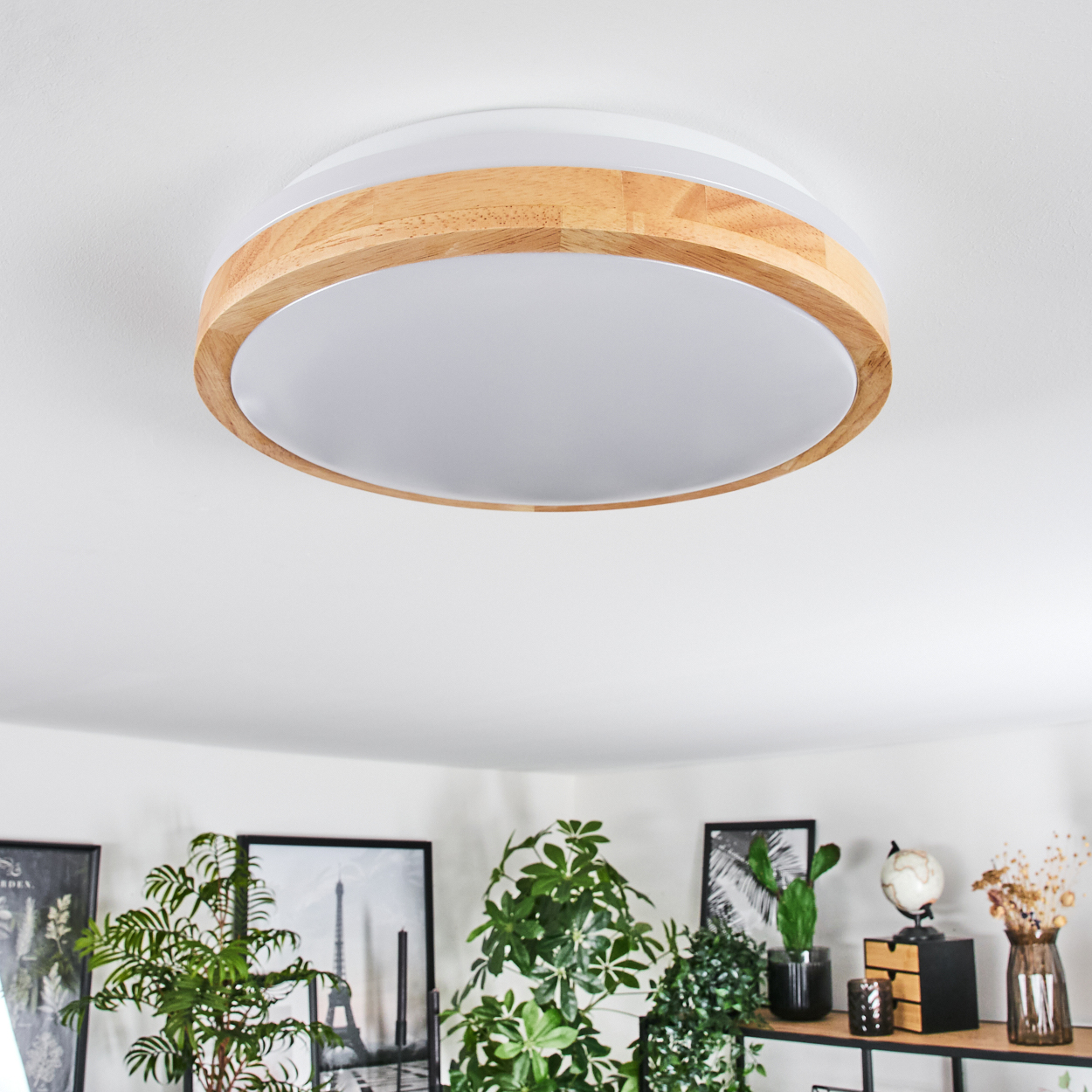 Eine runde Deckenlampe mit Holzrahmen und weißem Schirm. Im Hintergrund Pflanzen, Bilder und ein Regal mit Dekoration.