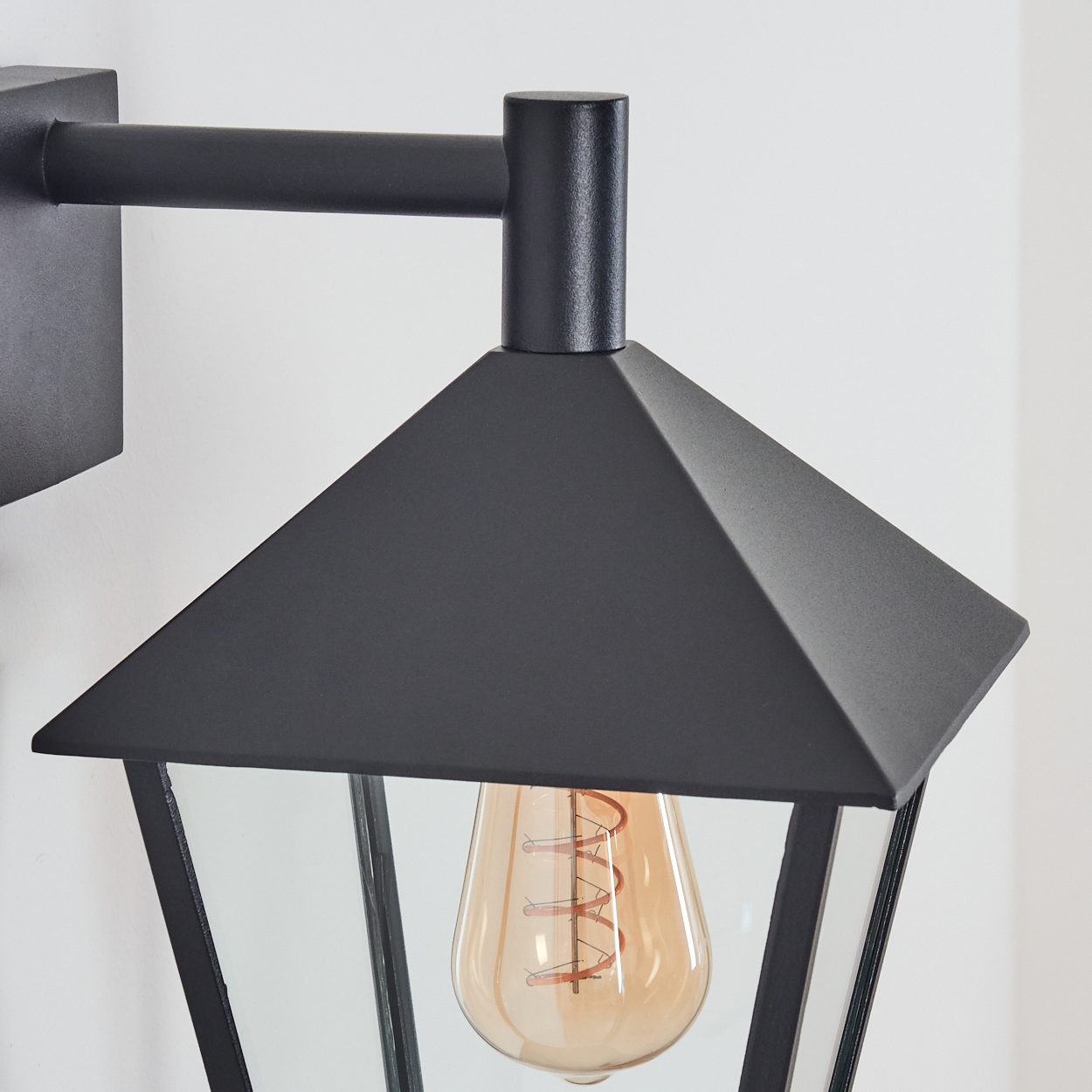 Eine schwarze Außenlampe mit Glasgehäuse und sichtbarer Glühbirne ist an einer weißen Wand montiert.