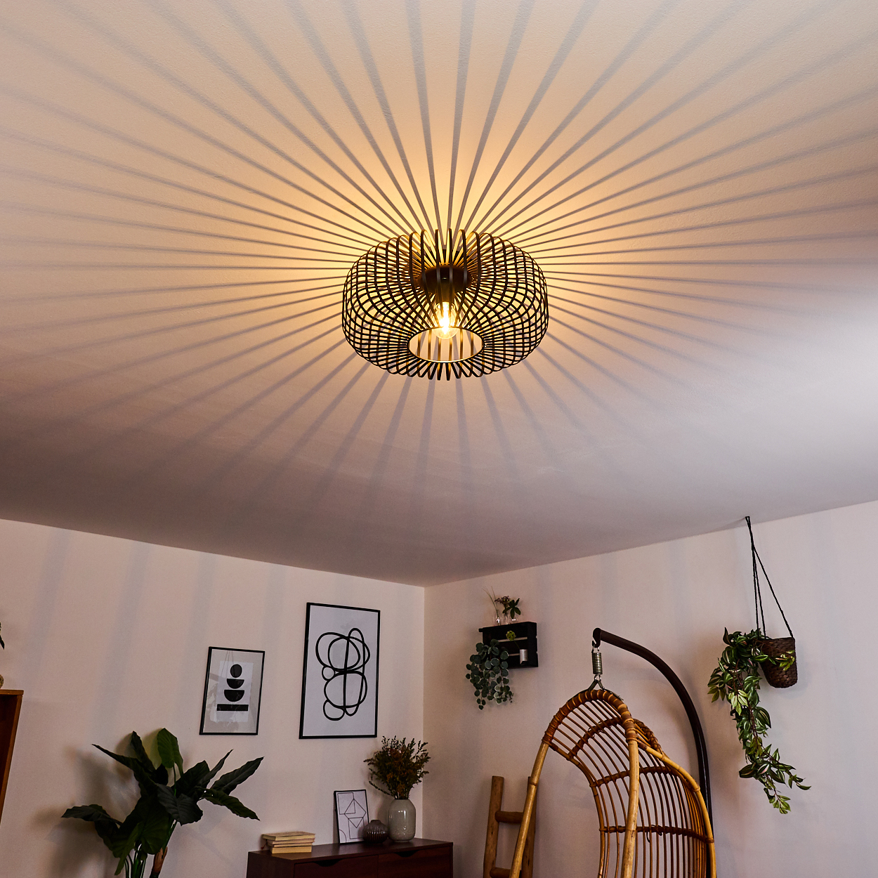 Eine schwarze, kreisförmige Deckenlampe wirft einen radialen Schatten. Innenraum mit Pflanzen, Kunst und einem hängenden Korbstuhl.