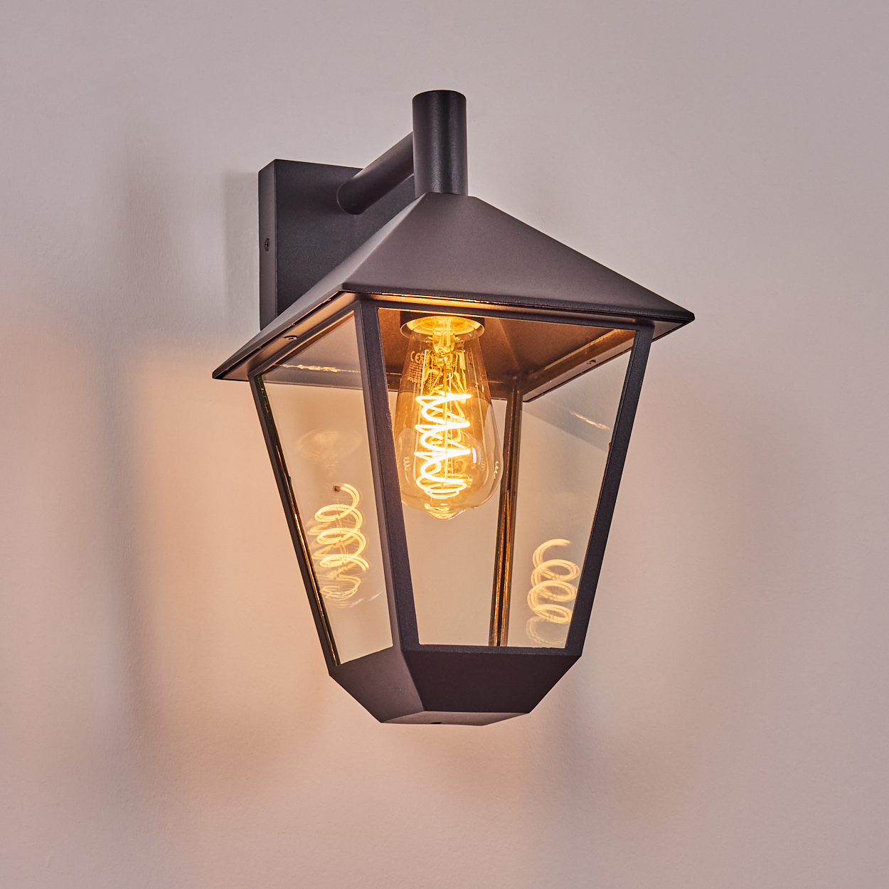 Schwarze Außenlaterne mit leuchtender Wendelglühbirne im Inneren, an einer hellen Wand montiert. Die Glasscheiben reflektieren das Licht.