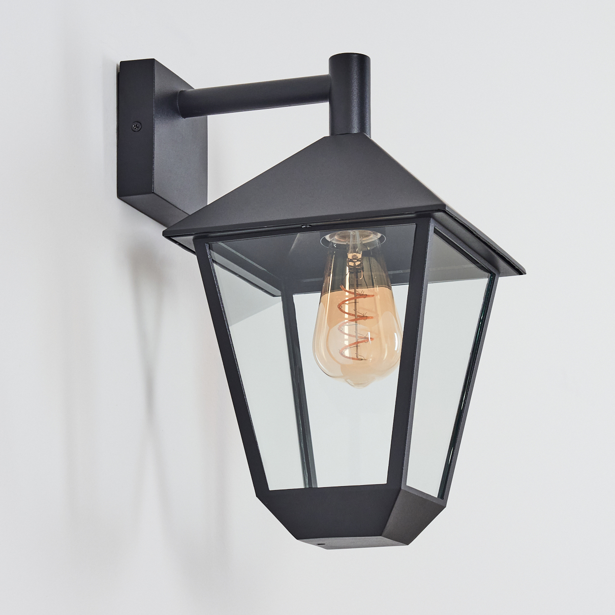 Eine schwarze Außenwandleuchte ist an einer weißen Wand montiert. Sie hat ein Glasgehäuse mit einer Edison-Glühbirne.