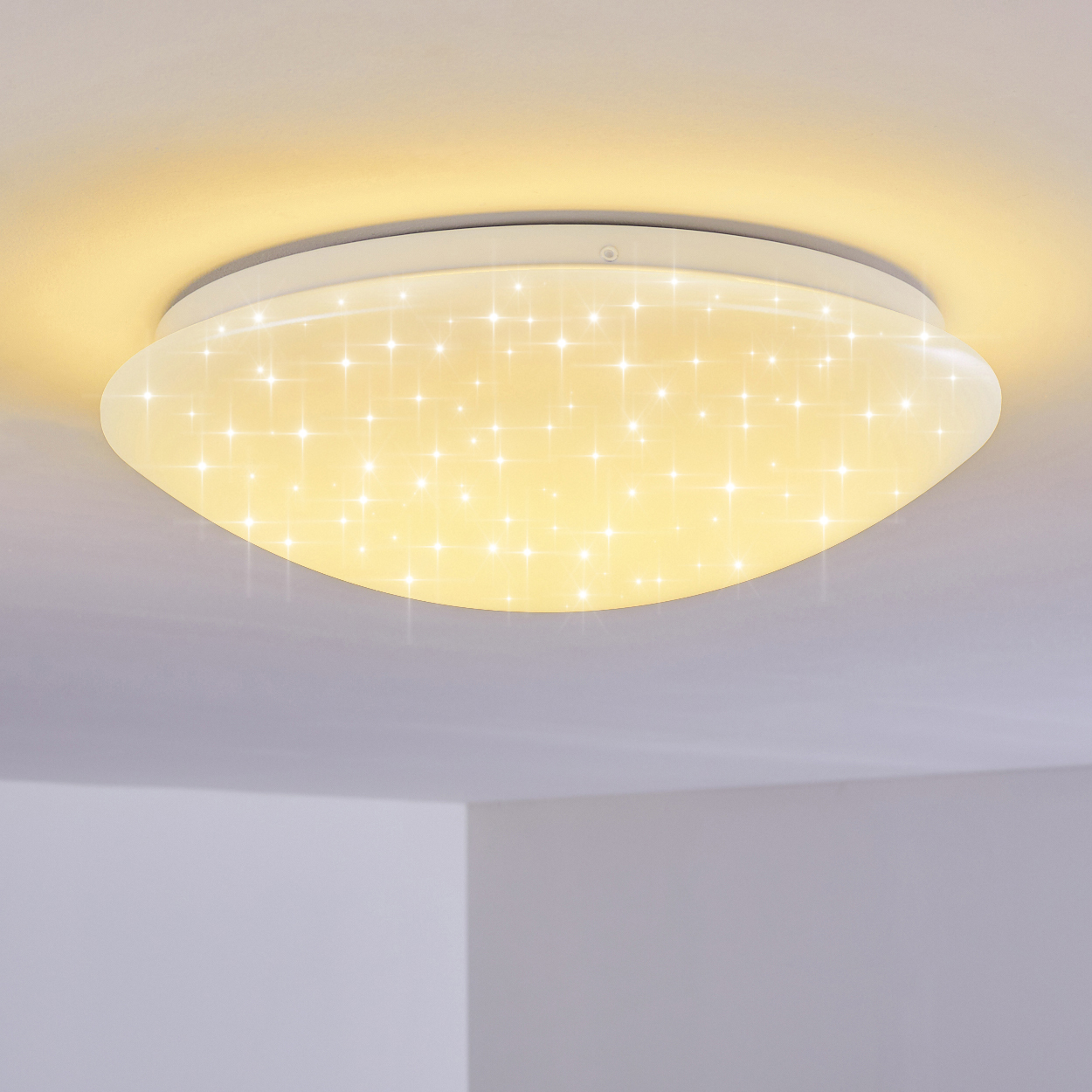 Eine Deckenleuchte mit sternförmigem Muster. Das Licht ist rund und strahlt warm. Die umgebende Decke ist weiß.