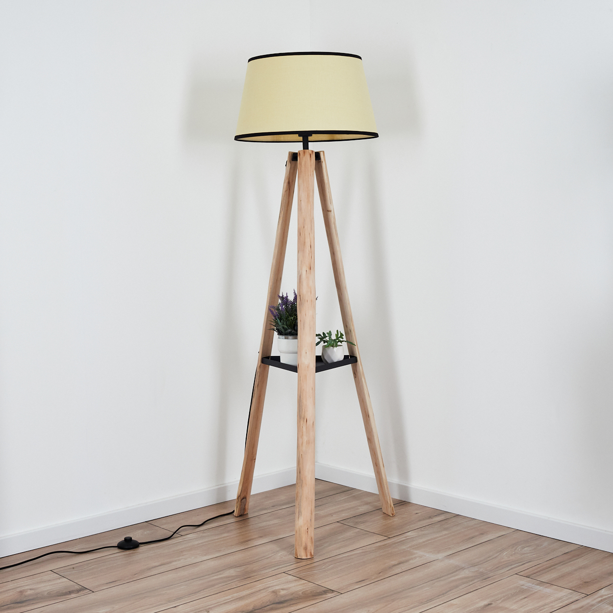 Eine hölzerne Stativ-Stehlampe mit cremefarbenem Schirm. Sie hat ein Regal mit Pflanzen. Der Hintergrund ist eine weiße Wand und ein Holzboden.