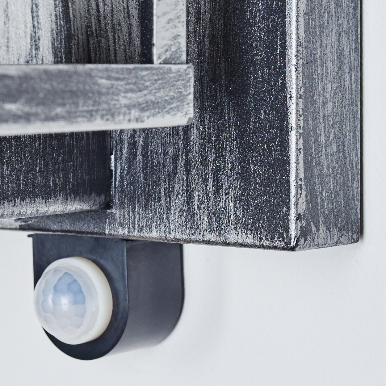 Nahaufnahme einer metallisch grauen Wandleuchte mit Bewegungssensor. Die Lampe ist an einer weißen Wand montiert. Der Sensor ist weiß und kuppelförmig.