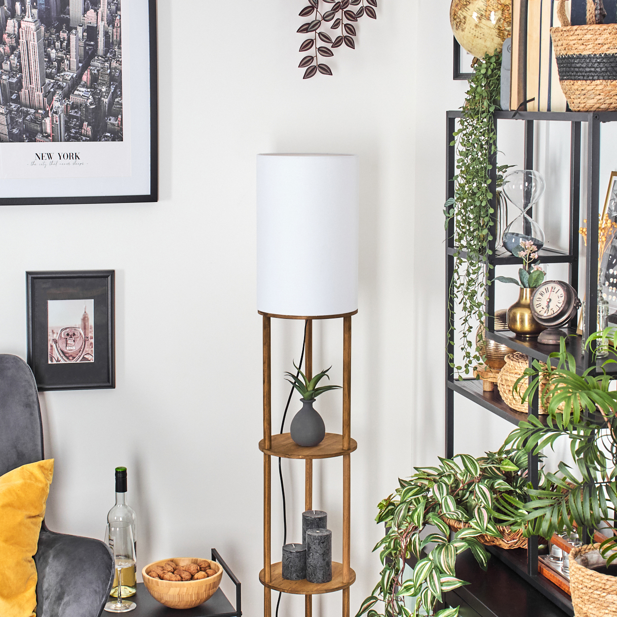 Eine Stehlampe mit Regalen hält eine Pflanze, Kerzen und eine Vase. Im Hintergrund befindet sich ein Bücherregal. An der Wand befindet sich ein gerahmtes New York-Foto.