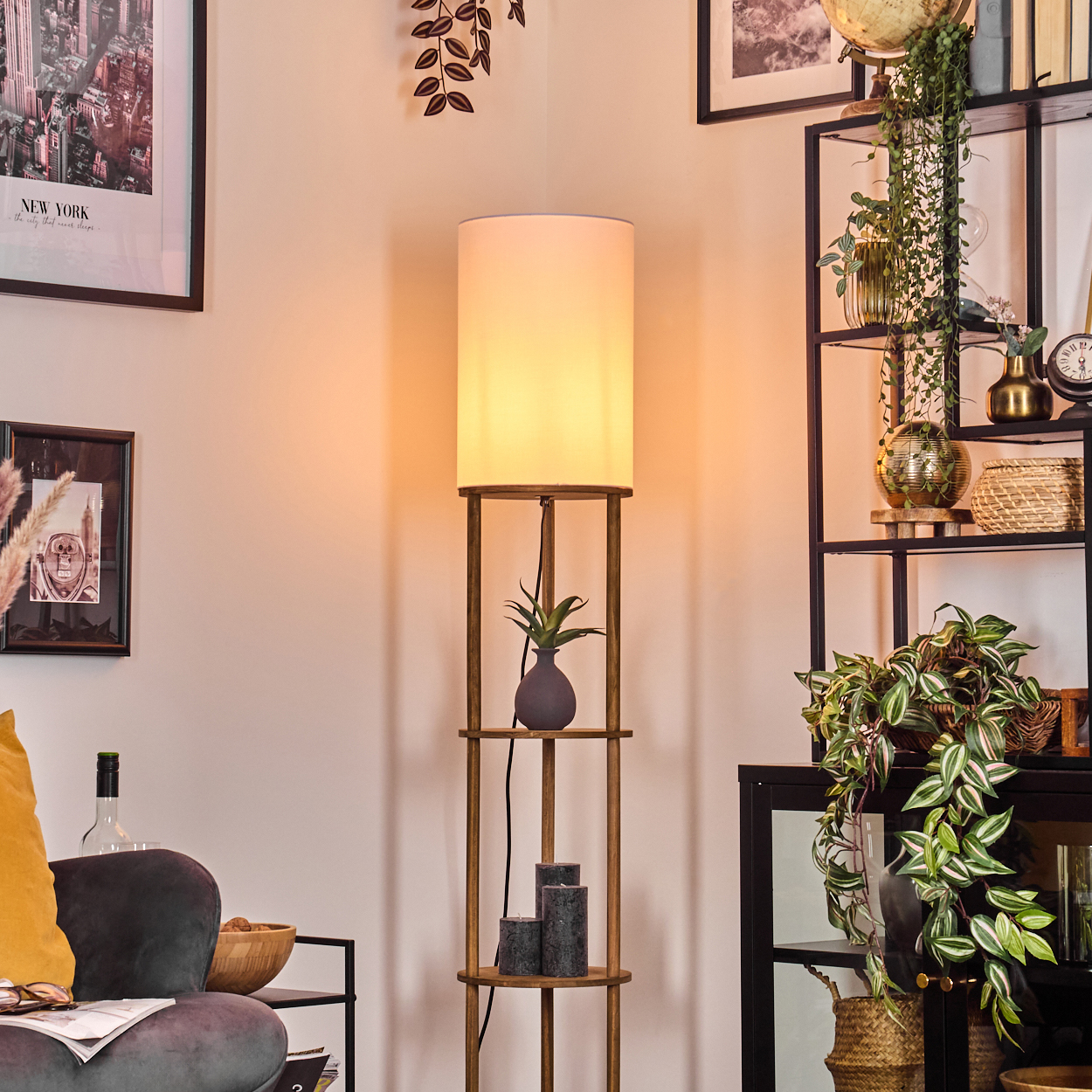 Eine Holzlampe mit weißem Schirm steht in einem Raum. Regale mit Pflanzen und Dekor sind sichtbar. Eine Couch mit gelbem Kissen steht in der Ecke.
