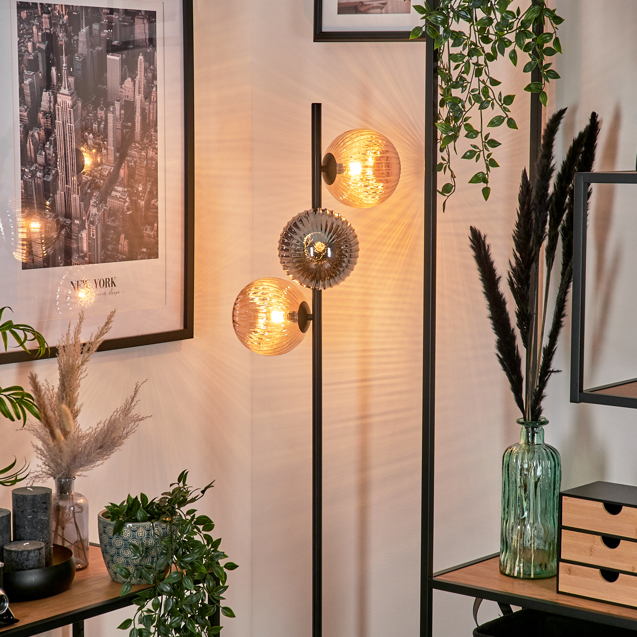 Ein modernes Interieur. Eine Stehlampe mit drei leuchtenden Kugeln. Ein New York-Druck und eine Vase mit schwarzen Schilf sind sichtbar.