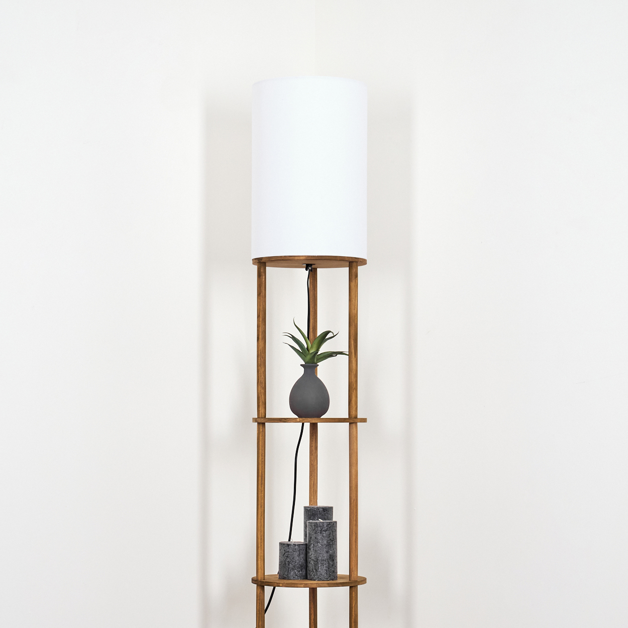 Eine hölzerne Stehlampe mit weißem Lampenschirm. Sie hat Regale mit einer Pflanze, einer Vase und Kerzen vor einer weißen Wand.