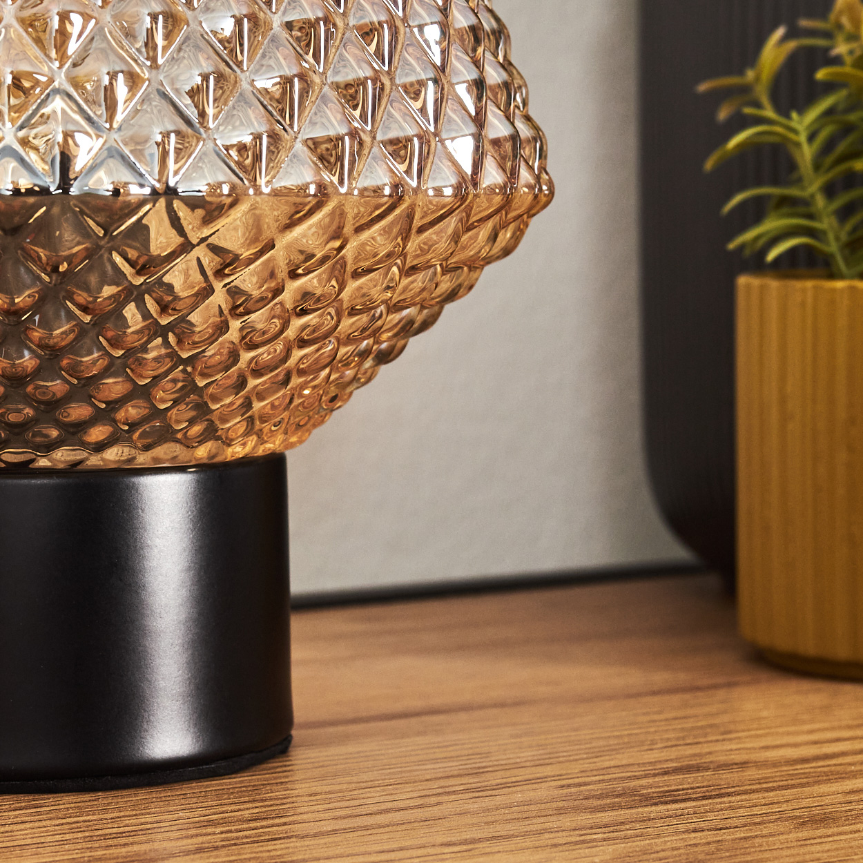 Detailaufnahme einer dekorativen Lampe mit strukturiertem Glasschirm. Im Hintergrund eine Pflanze in einem gelben Topf.