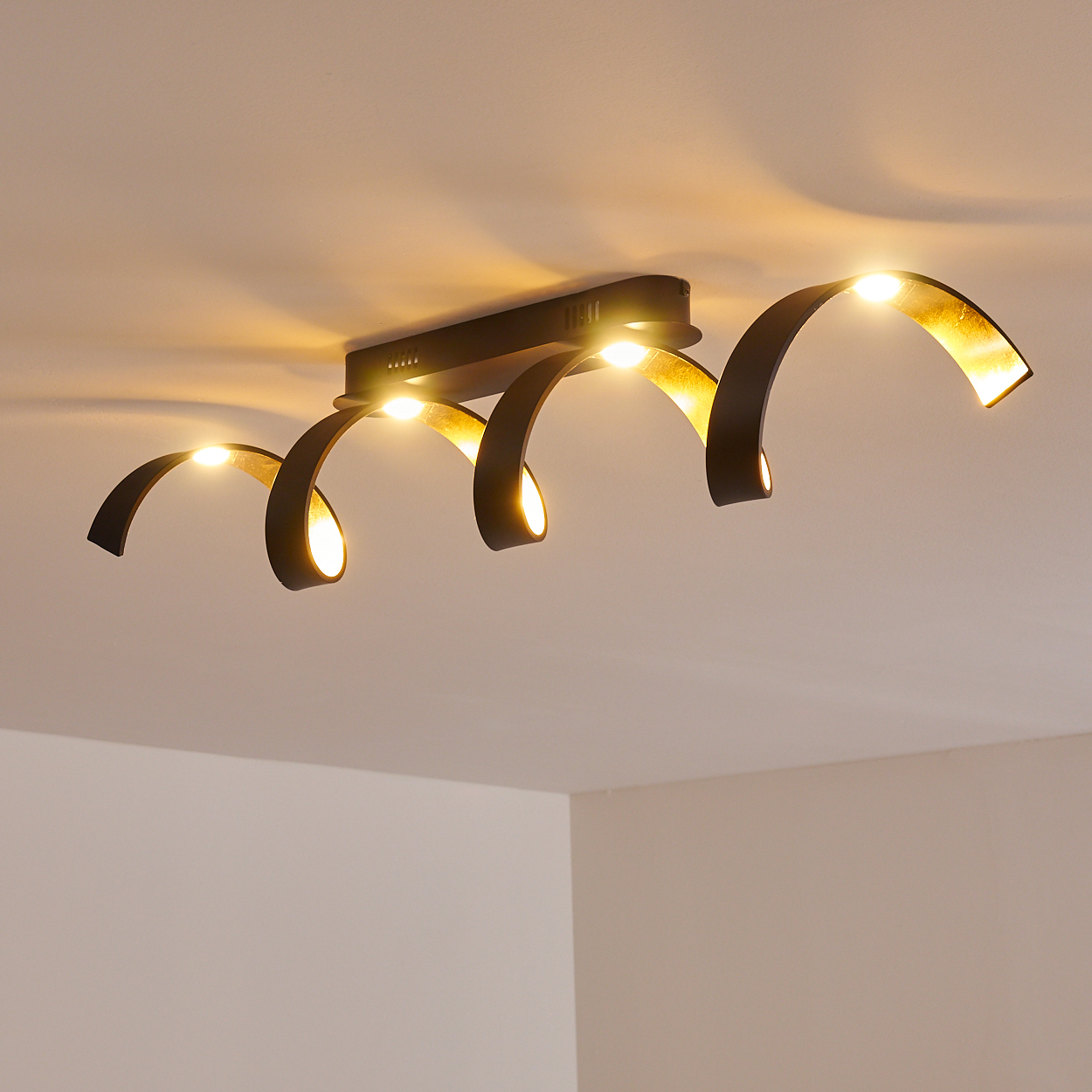 Eine moderne Deckenleuchte mit fünf gebogenen, schwarzen Armen. Jeder Arm hält ein warmes, leuchtendes Licht. Die Leuchte ist an einer weißen Decke montiert.