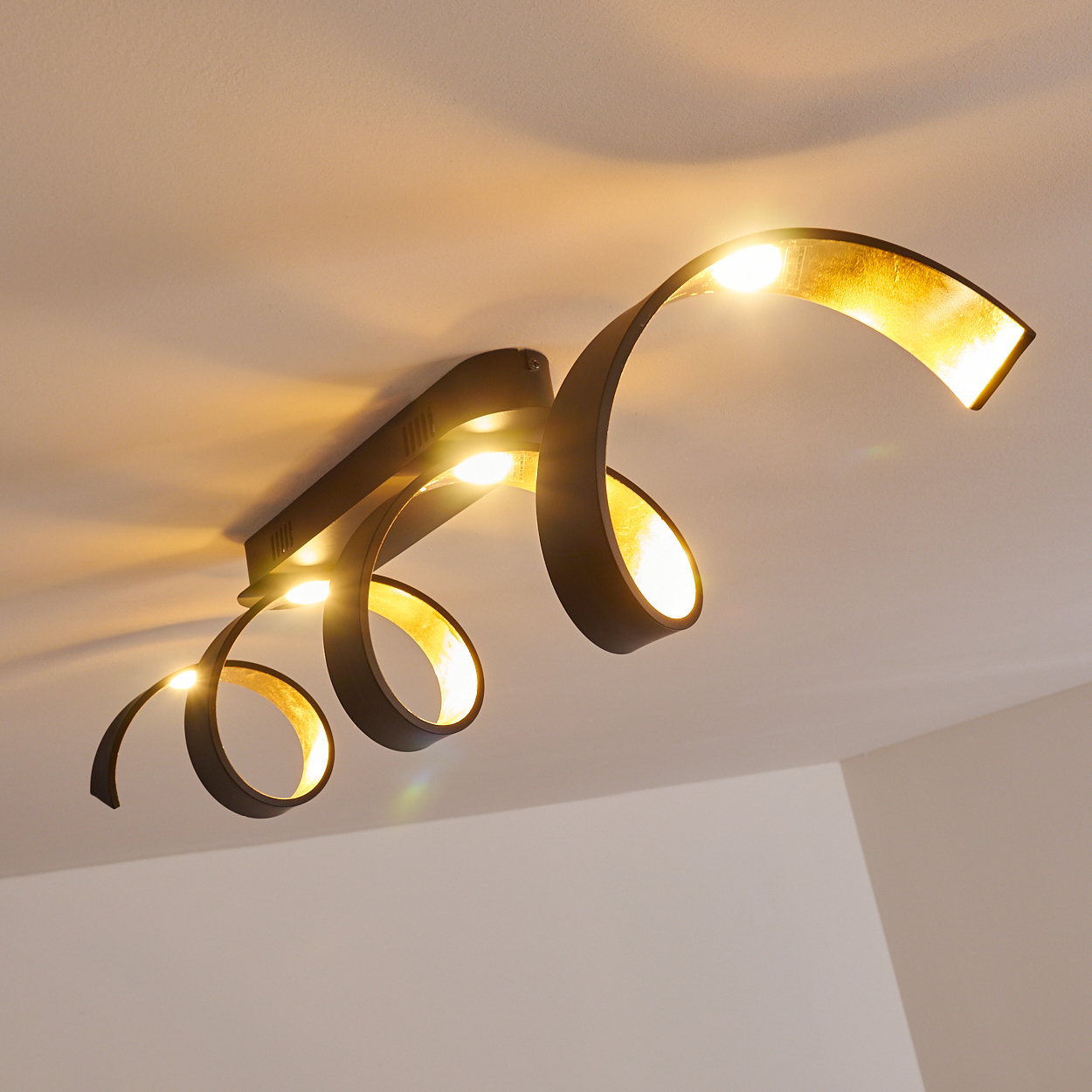 Moderne Deckenleuchte mit schwarzen, wirbelnden Armen und Blattgold-Interieur. Das Licht strahlt warm auf einer weißen Decke.