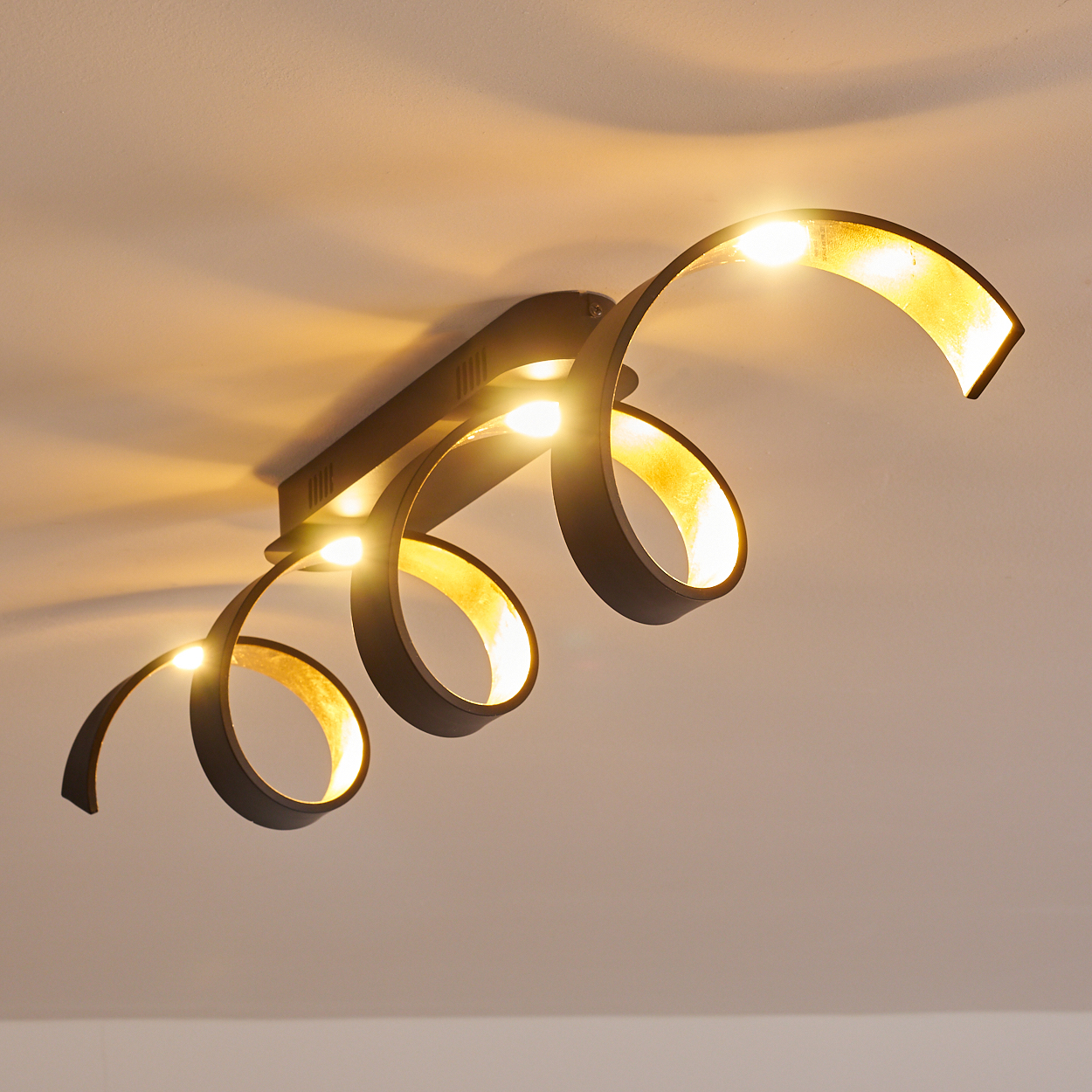 Eine moderne Deckenleuchte mit einem wirbelnden Design. Sie hat einen schwarzen Rahmen und ein goldenes Innere mit mehreren hellen Lichtern.