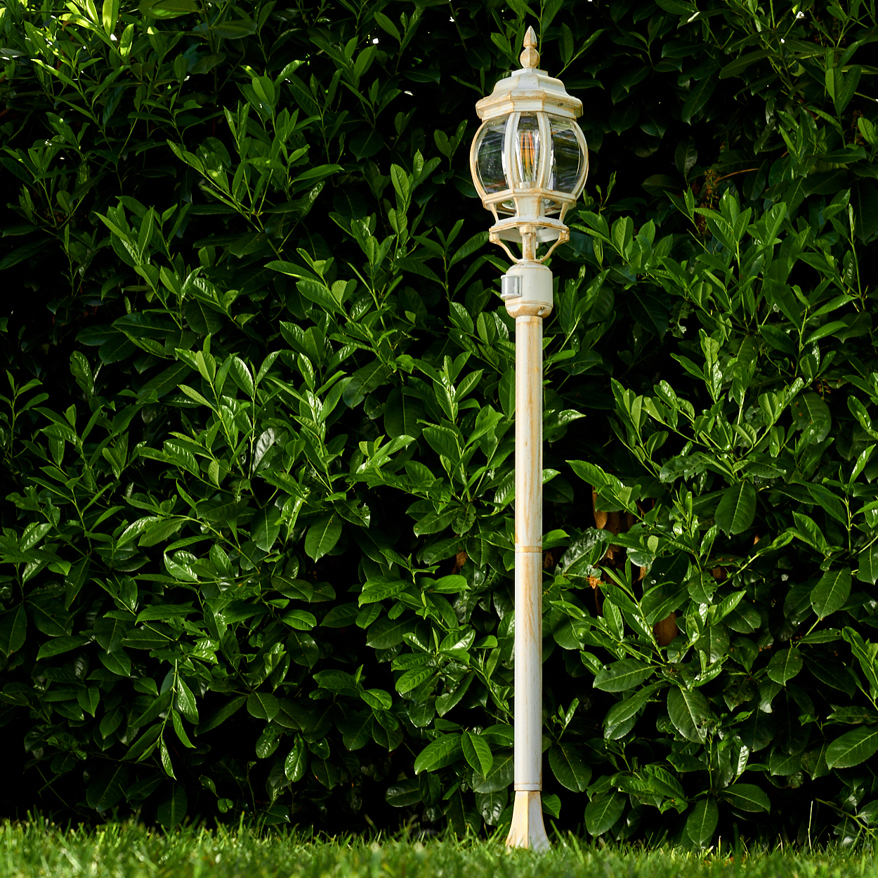 Eine weiße, verzierte Straßenlaterne steht auf grünem Gras vor einer dichten grünen Hecke. Die Lampe hat eine Glaskugel.