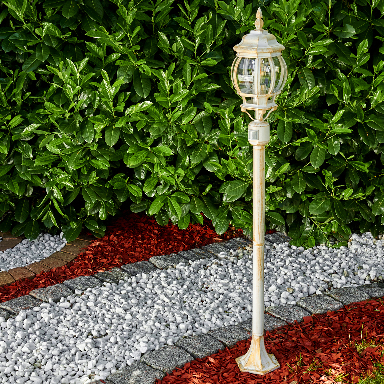 Eine weiße Straßenlaterne steht in einem Garten. Dahinter befindet sich ein üppiger grüner Busch. Ein Weg besteht aus weißen Steinen, roter Mulche und Bordsteinen.