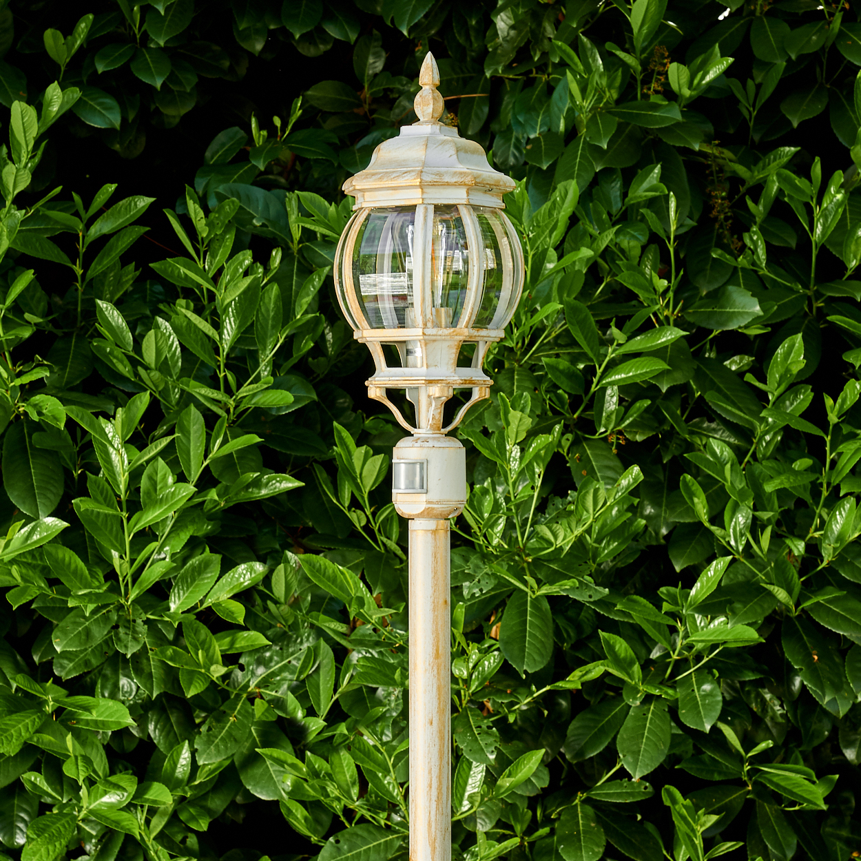 Eine weiße antike Straßenlaterne vor einem grünen, belaubten Hintergrund. Die Lampe hat eine klare Glaskugel und eine dekorative Spitze.