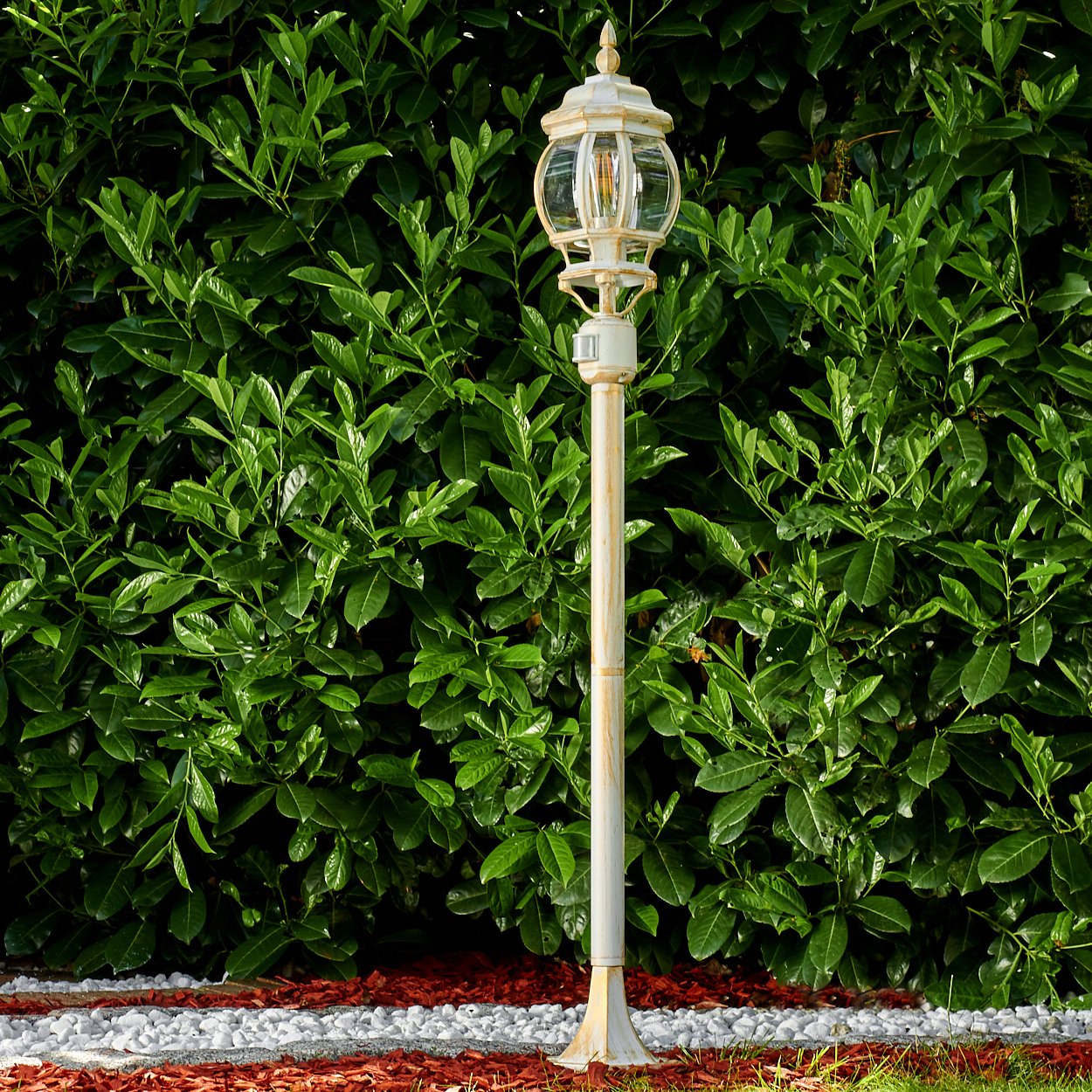 Eine weiße Laterne steht vor einer grünen Hecke. Roter Mulch, weiße Steine und Gras bilden die Basis. Die Lampe hat ein Glasgehäuse.