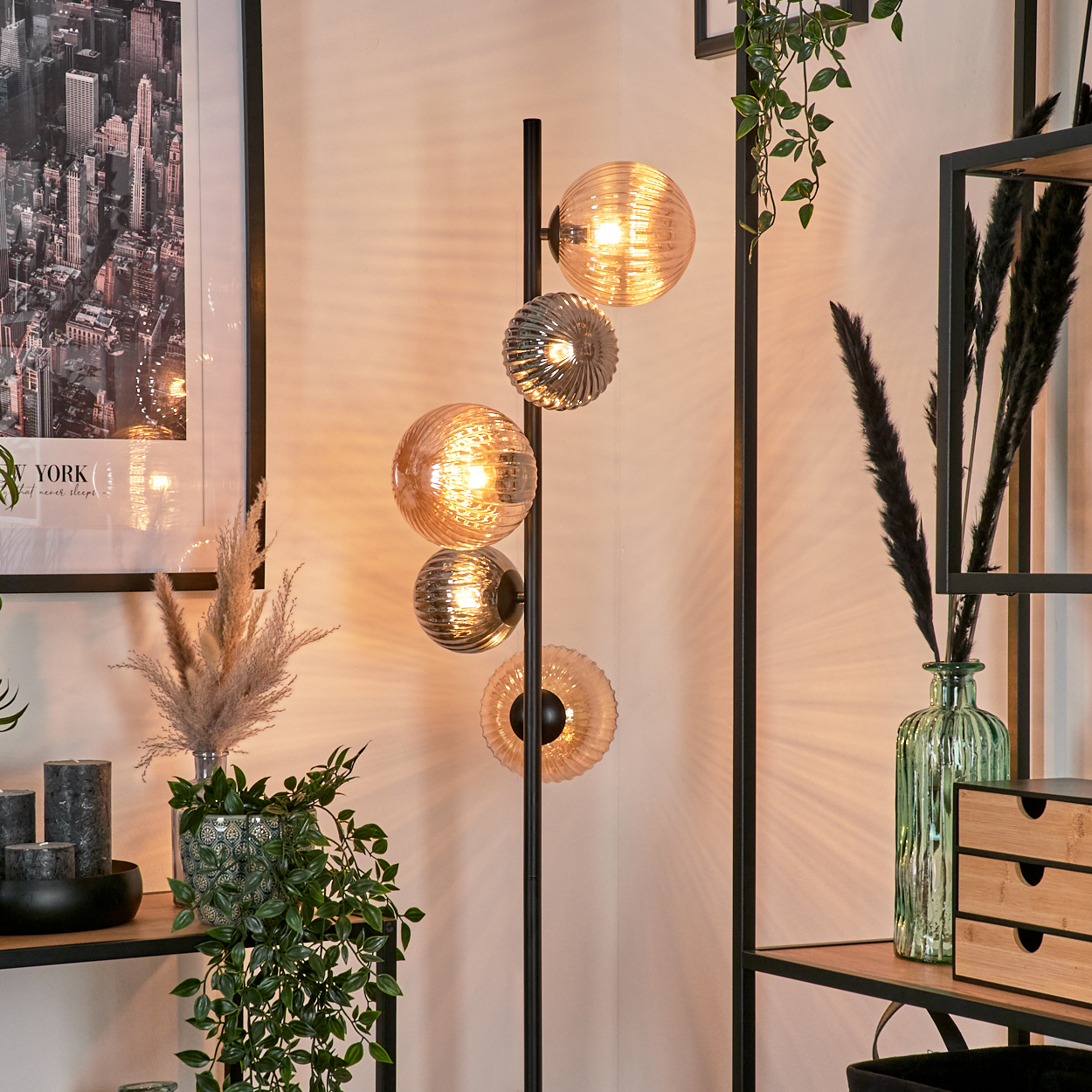 Eine stilvolle Stehlampe mit Glaskugelschirmen. Sie steht neben einem Regal mit Pflanzen und Dekorationsartikeln, darunter ein Stadtbild.