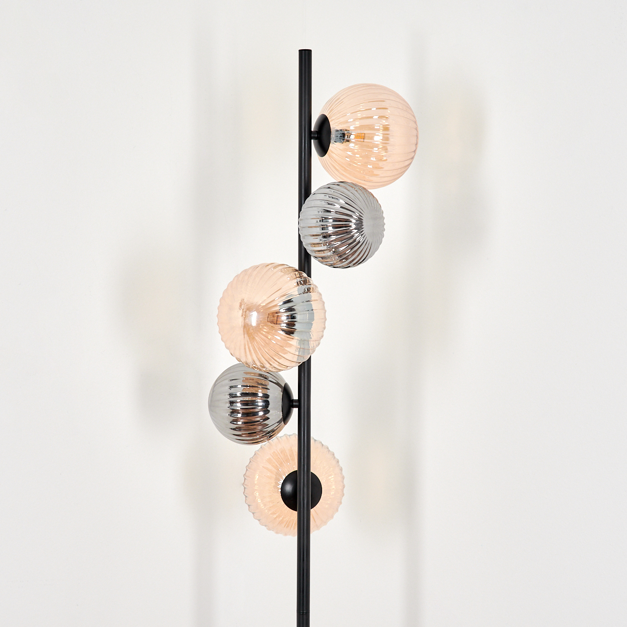 Eine schwarze Stehlampe mit fünf kugelförmigen Schirmen in Pfirsich und Silber. Die Schirme sind gerippt und der Hintergrund ist eine schlichte weiße Wand.
