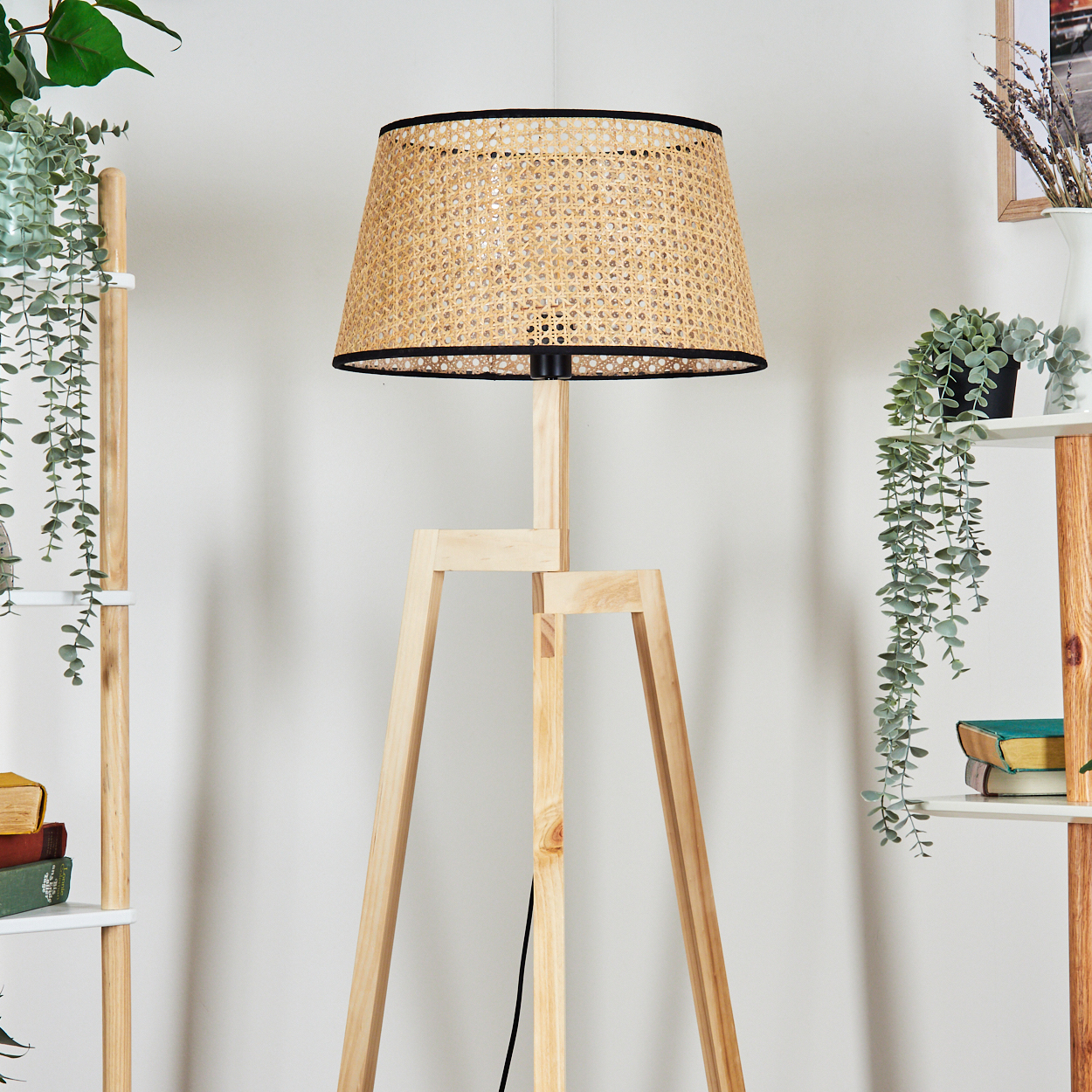 Eine hölzerne Stativlampe mit gewebtem Schirm steht vor einer weißen Wand. Grüne Pflanzen und Regale sind auf beiden Seiten.